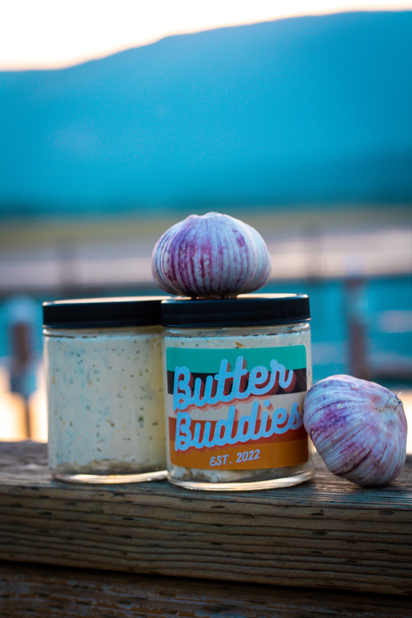 Butter Buddies Garlic Butter with whole cloves of garlic on top and all around on a dock, during sunset.