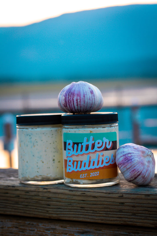 Butter Buddies Garlic Butter with whole cloves of garlic on top and all around on a dock, during sunset. 